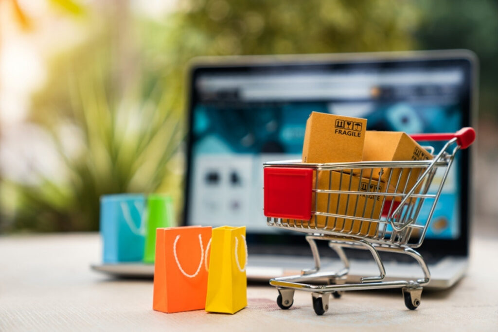 Einkaufswagen mit Paketen und bunten Papiertüten vor einem Laptop, auf dem ein Onlineshop geöffnet ist